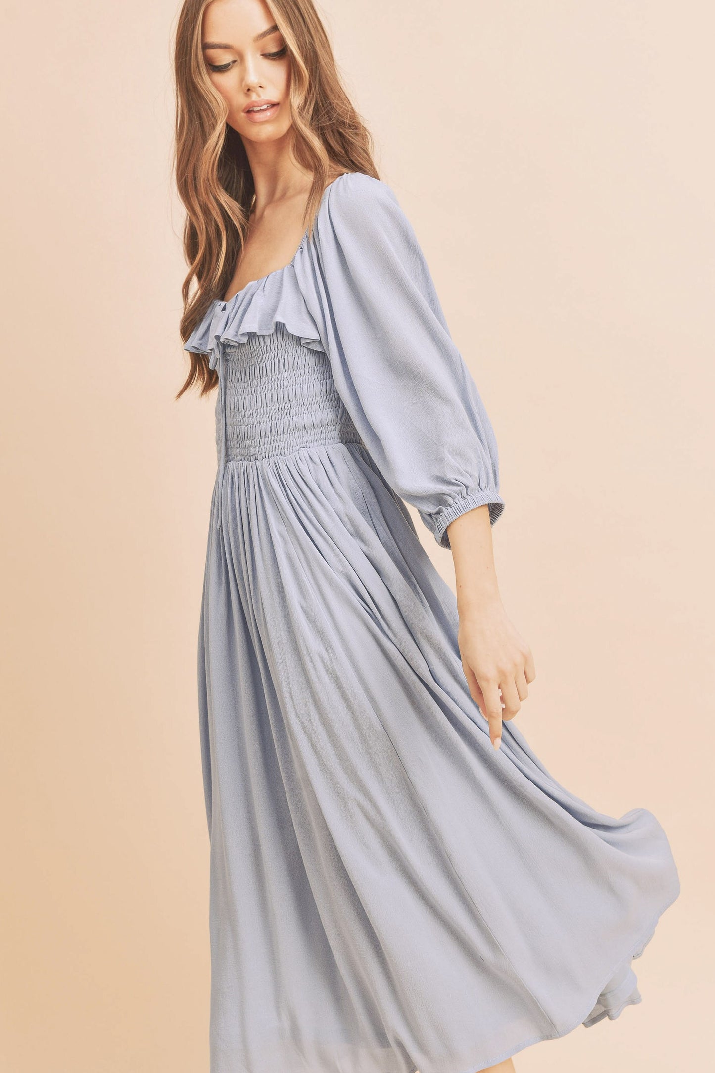 The Geri Dress boasts an eternally flattering shape, featuring a charmingly tied ruffled neckline, a smocked bodice, a lined skirt, and voluminous sleeves.